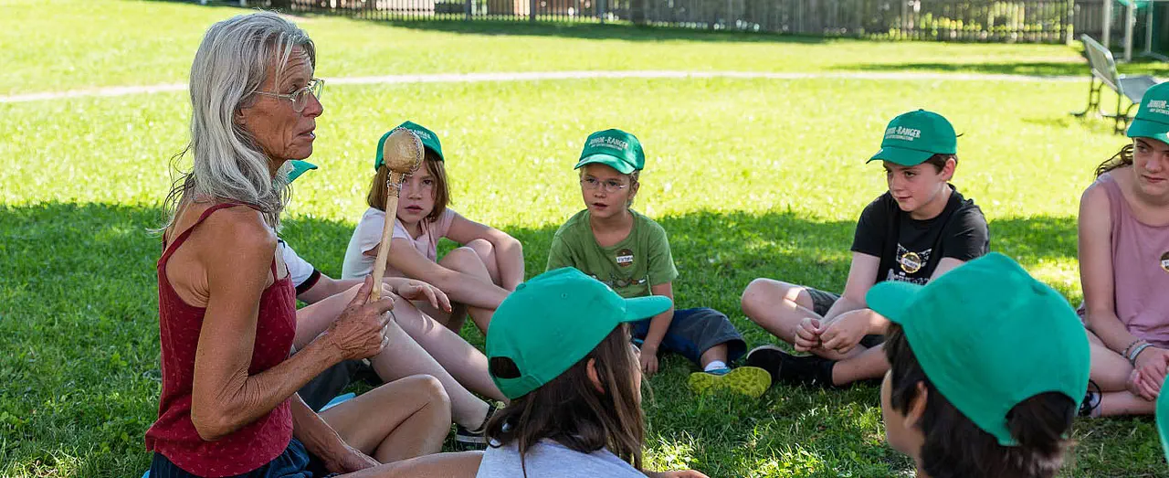 Auf diesem Foto erzählt etwas den im Kreis auf einer Wiese sitzenden Kindern eine Natur- und Landschaftsführerin.