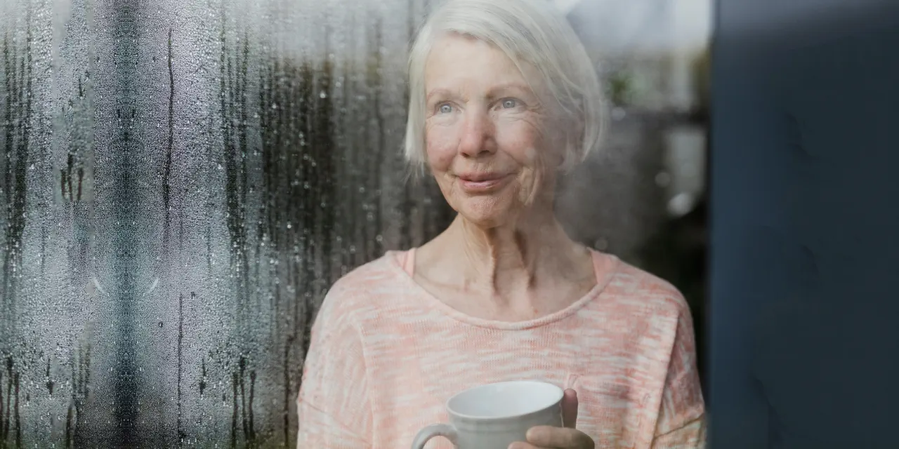 Ältere Frau mit einer Tasse vor einem nassen Fenster