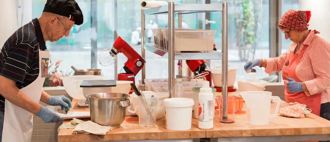 Zwei ältere Mitarbeiter von Kuchentratsch stehen sich gegenüber an einem großen Tisch und arbeiten mit Teig.